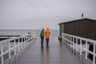 Rear view of men walking on wet pier against sea and sky during rainy season