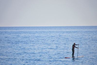 Man standing on paddleboard in sea against clear sky