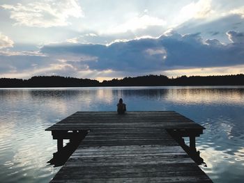 Woman sitting on pier over lake against sky during sunset