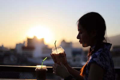 Side view of woman having drink in city against clear sky during sunset