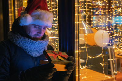 Man wearing santa hat opening gift box by window