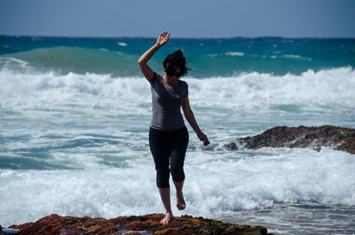 Smiling young woman with arms raised standing on rock against sea