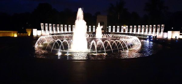 Illuminated fountain by river against sky at night