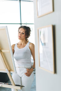 Thinking woman artist in white clothes with paint brushes in front of easel in modern art gallery