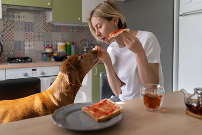 Middle aged mature woman stroking and treating dog with sandwich sits in kitchen during breakfast