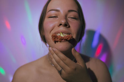 Close-up of smiling young woman eating fig against colorful lights on wall