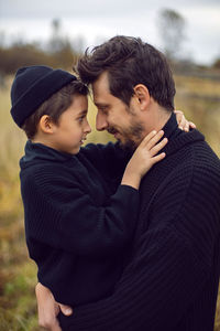 Portrait father and son in black clothes stand in a field with an old wooden horse fence in the fall