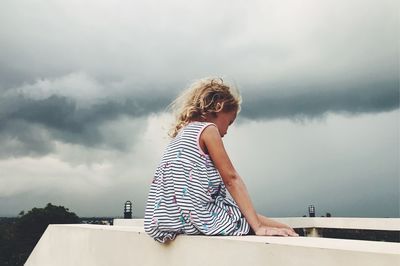 Girl sitting on retaining wall against sky