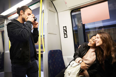 Young man photographing happy female friends sitting in train