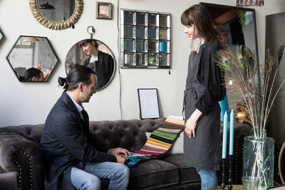 Young female owner looking at mature male customer choosing from fabric swatch while sitting on sofa at store