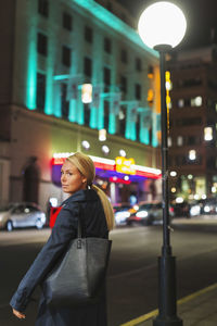 Side view of woman standing on sidewalk in city during night