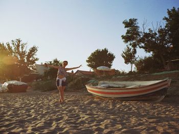 Full length of woman with arms outstretched standing by boat at sandy beach