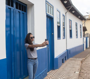 Smiling young woman taking selfie while standing by wall