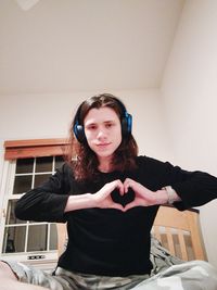 Portrait of young woman making heart shape with hands while listening to music at home