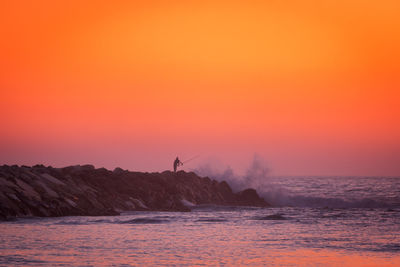 Fisherman silhouette fishing with waves crashing on a stone pier at sunset, in portugal