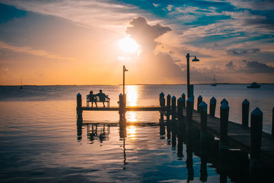 Silhouette friends sitting on bench at pier over sea against sky during sunset