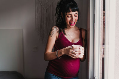 Smiling pregnant woman with mug standing by window at home