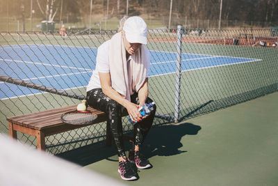 Full length of tired woman sitting on bench against net at tennis court