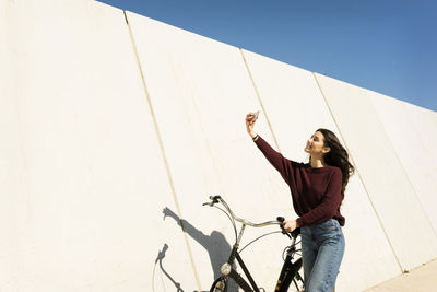Woman with bicycle taking selfie though smart phone by wall