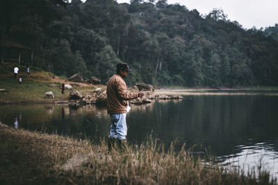 Rear view of man standing by lake against trees