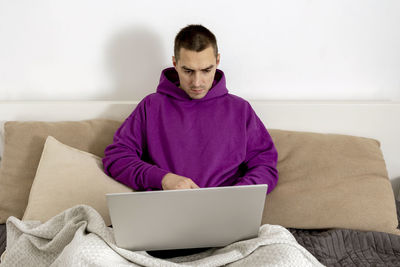 Young caucasian man with violet hoodie sitting on bed and holding laptop computer.