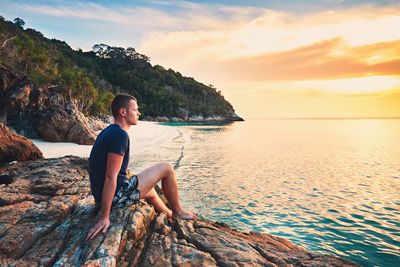 Young man relaxing on rock formation while looking at sea against sky during sunset