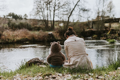 Rear view of mother and daughter sitting by lake in forest