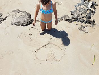 Girl wearing bikini while looking at heart shape on sand at beach during sunny day