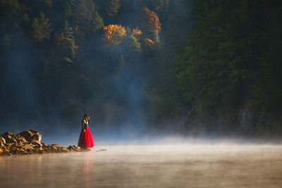 Woman standing at lakeshore in forest during foggy weather