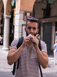 Portrait of young man eating while standing on street in city