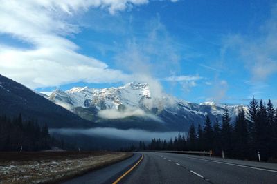 Empty road by snowcapped mountains against cloudy sky