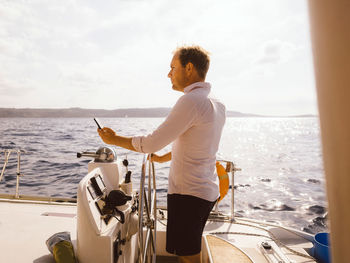 Side view of man holding mobile phone while sailing catamaran on sea against sky during sunny day