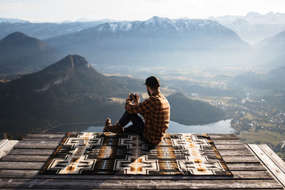 Back view of unrecognizable male tourist sitting on blanket at viewpoint and taking photo