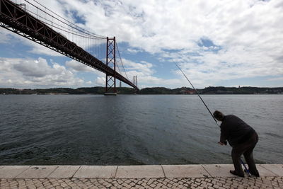 Rear view of man fishing while standing on jetty by 25 de abril bridge