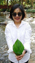 Portrait of cheerful woman holding leaf while standing at riverbank in forest