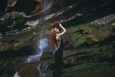Side view of beautiful redhead woman standing against waterfall
