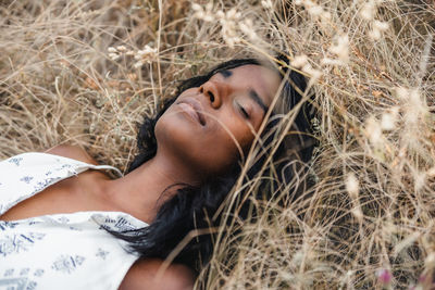 Mindful adult ethnic female with closed eyes and dark hair lying on dry grass in daytime