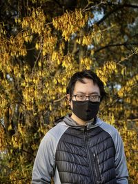 Portrait of young asian man in eyeglasses and face mask against flowering laburnum tree at sunset
