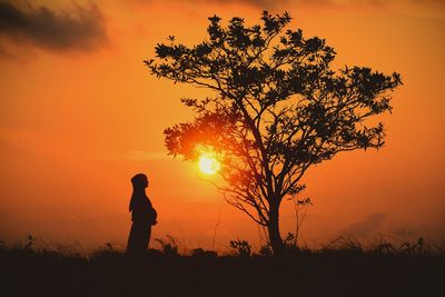 Silhouette pregnant woman standing on field by tree against orange sky