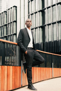 Full body positive african american manager with hand in pocket of suit leaning on fence and looking away on city street