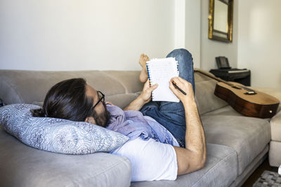 Back view of smart bearded man in casual wear lying on comfortable couch while taking notes in stylish living room