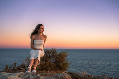 Young attractive female in stylish dress sitting on rocky cliff near waving sea against sundown sky 