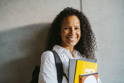 Portrait of smiling woman with curly hair holding book and backpack leaning on gray wall at college