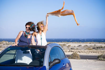 Happy female friends standing in car at beach against clear blue sky