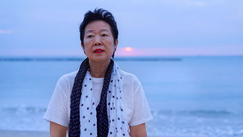 Thoughtful senior woman standing at beach against sky during sunset