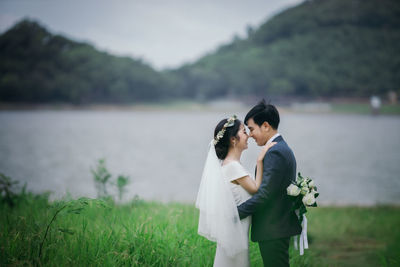 Married couple embracing amidst grass against lake