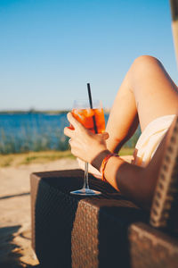 Low section of young woman holding drink while sitting on chair at beach against clear sky during sunny day