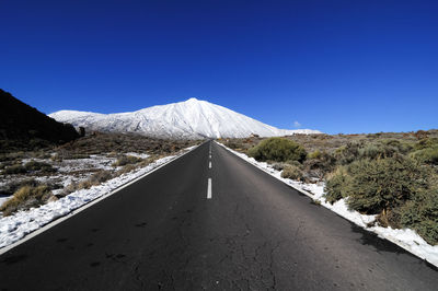 Road leading towards mountains against clear blue sky