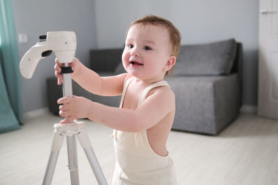 European child of 1 year in white jumpsuit with tripod for photo of house in bright real