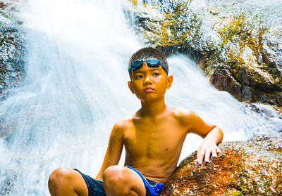 Shirtless boy sitting by rock against waterfall